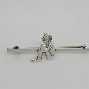 May include: A silver-toned metal brooch featuring a horizontal bar with a small, seated figure wearing a pointed hat attached to the center. The figure is depicted with knees pulled toward the chest, and the brooch includes a pin mechanism on the back.