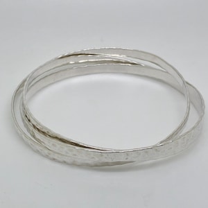 Puede incluir: Tres pulseras plateadas con textura martillada se muestran sobre un fondo blanco. Las pulseras están entrelazadas, creando un efecto de capas. El color plateado refleja la luz, resaltando la superficie texturizada de la joyería.