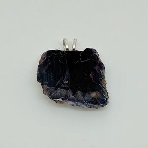 May include: A raw, dark purple and black stone pendant with an irregular, jagged shape and a polished surface. The stone features a silver-toned metal bail attached to the top edge, allowing it to be suspended from a chain or cord for jewelry.