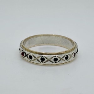 May include: Silver ring with a repeating pattern of black and red gemstones set in a diamond-shaped design. The ring has a gold-colored band on the inside and a silver band on the outside. The ring is displayed on a white background.