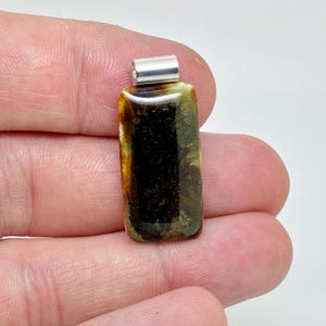 May include: A rectangular pendant featuring a glossy, dark brown and amber-colored surface with swirling patterns. The pendant is topped with a polished silver-toned metal bail, providing a secure attachment point for a necklace chain or cord.