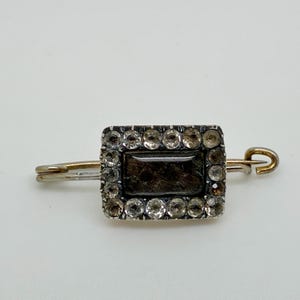 May include: Antique rectangular brooch with a dark center stone surrounded by clear, round accent stones. The brooch has a silver-colored frame and a gold-colored pin and clasp. The jewelry is set against a white background.