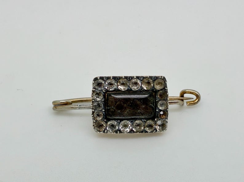 May include: Antique rectangular brooch with a dark center stone surrounded by clear, round accent stones. The brooch has a silver-colored frame and a gold-colored pin and clasp. The jewelry is set against a white background.