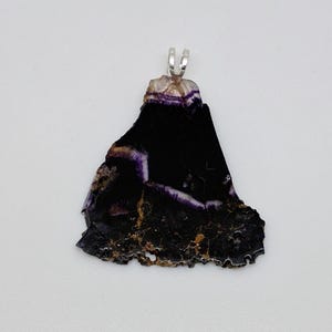 May include: A pendant featuring a dark, irregularly shaped stone with a silver bail. The stone is primarily black with streaks of purple and hints of gold. The pendant has a rough, natural texture.