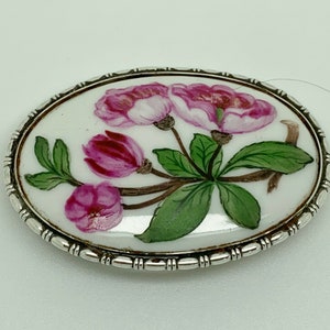Pode incluir: Um broche oval de porcelana com uma borda prateada. O broche apresenta um desenho pintado à mão de flores cor-de-rosa com folhas e caules verdes.