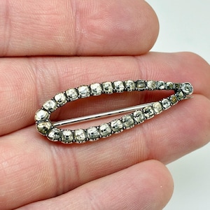 May include: A vintage silver-toned brooch in an oval shape, encrusted with numerous clear, sparkling stones. The brooch features a pin on the back for secure fastening. The stones are set closely together, creating a continuous line of brilliance.