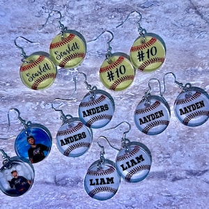 May include: A set of 10 pairs of baseball-themed earrings. The earrings feature different designs, including names, numbers, and photos. The earrings are made of clear resin and have silver hooks.