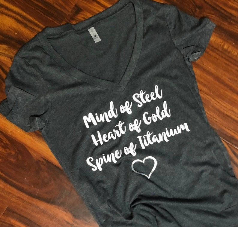 May include: A dark grey V-neck t-shirt with the text "Mind of Steel, Heart of Gold, Spine of Titanium" in white lettering and a white heart.