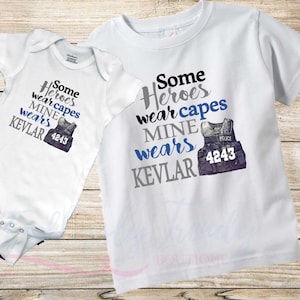 May include: Two white t-shirts with a graphic design of a police vest and the text "Some heroes wear capes MINE wears KEVLAR 4243".