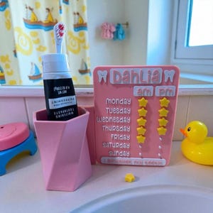 May include: A pink geometric toothbrush holder with a tube of peppermint toothpaste and a toothbrush. A pink daily teeth brushing chart with the name "Dahlia" and star-shaped markers sits next to a yellow rubber duck.