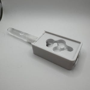 May include: A white rectangular plastic mold featuring a central cutout with a complex, organic shape, attached to a clear, cylindrical plastic handle. The mold has a smooth, matte finish and is designed for casting or shaping materials.