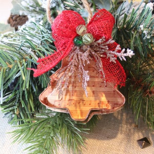 Puede incluir: Un adorno navideño de metal marrón en forma de campana, decorado con notas musicales, está adornado con un lazo de arpillera rojo con cuentas verdes y doradas. El adorno está rodeado de ramas de pino verdes.