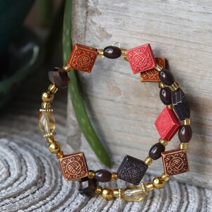 Könnte beinhalten: Ein braunes, schwarzes und rotes Perlenarmband mit quadratischen Anhängern mit chinesischen Schriftzeichen. Das Armband besteht aus mehreren Perlensträngen und ist für das Tragen am Handgelenk gedacht.