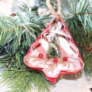 May include: A red and white striped Christmas tree ornament made from a cookie cutter. The ornament is decorated with a small green bottle brush tree, red beads, and gold glitter.