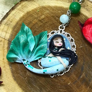 Peut inclure: Un collier en argent avec un pendentif en forme de sirène bleu et vert. La sirène a de longs cheveux noirs et est assise sur une queue bleue et verte. Le collier est composé de perles argentées et d'une perle verte et bleue au sommet.