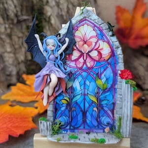 Peut inclure: Une figurine de fée miniature aux cheveux bleus et aux ailes noires, à côté d'un vitrail. La fenêtre présente un motif floral vibrant dans les tons de rose, de bleu et de violet. La scène est posée sur un petit chevalet en bois, avec des feuilles d'automne en arrière-plan.