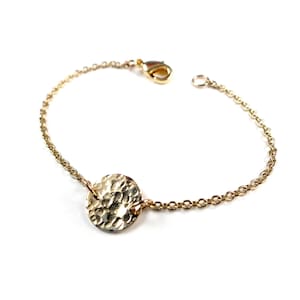 Puede incluir: Un brazalete dorado con un colgante de disco martillado. El brazalete presenta una delicada cadena y un cierre de mosquetón. El colgante tiene una superficie texturizada.