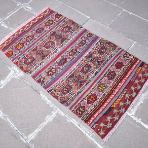May include: A red, blue, and yellow patterned rug with fringe on the edges. The rug has a geometric design and is laid out on a gray tiled floor.