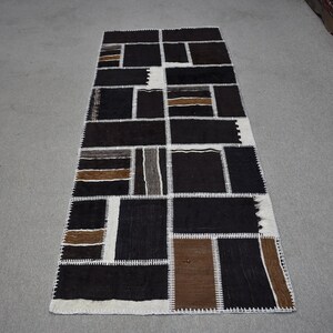 Op de afbeelding: Een lang, rechthoekig tapijt met een patchwork-ontwerp. Het tapijt is gemaakt van verschillende gekleurde stoffen, waaronder zwart, bruin en wit. Het tapijt heeft een witte rand en is aan elkaar gestikt met wit garen.
