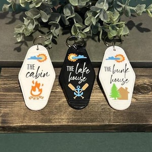 May include: Three retro-style keyrings with the text "The Cabin", "The Lake House", and "The Bunk House". Each keyring has a different design, including a campfire, an anchor and oars, and a tree and house. The keyrings are white and black.