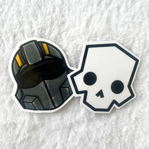 May include: Two stickers: one of a gray and black helmet with yellow accents, and another of a white skull with black eye sockets and a black outline. The stickers are on a white, textured surface.