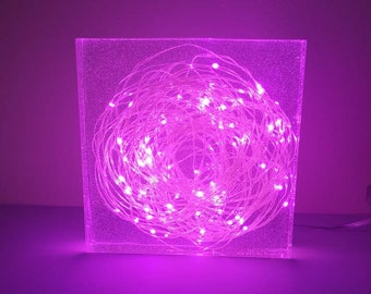 May include: A square clear glass lamp with a pink light inside. The light is made of a string of small white lights that are tangled together in a ball shape.