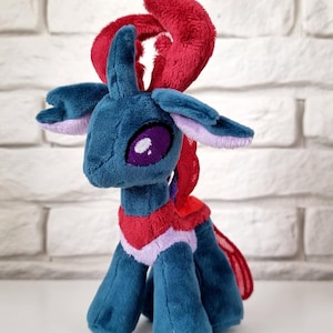 Könnte beinhalten: Ein Plüschtier, das eine Kreatur mit tealfarbenem Fell, großen lila Augen und roten und rosa Akzenten darstellt. Das Spielzeug hat ein stilisiertes Design mit langem Hals, kleinen Ohren und einer flauschigen roten Mähne.