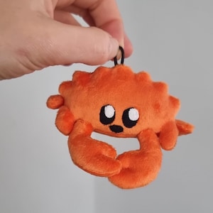 May include: An orange plush crab keychain with black eyes and a black mouth. The crab has two large claws and a textured shell. The keychain is held by a black loop.