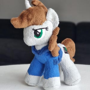 Puede incluir: Un peluche de pony con cuerpo blanco, pelo castaño y un atuendo azul. El pony tiene ojos verdes y un cuerno blanco. El juguete está hecho de material suave y difuso, y está sobre una superficie blanca.