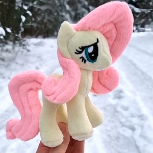 May include: A soft toy pony, featuring a light yellow body and a pink mane and tail. The pony has large eyes with blue and black detailing. The background shows a snowy outdoor scene.
