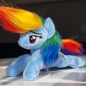 Könnte beinhalten: Ein hellblaues Plüschpferd mit Regenbogenmähne und -schwanz. Das Pony hat gestickte Augen und ein Schönheitsfleck auf der Flanke. Das Spielzeug liegt auf einer weißen Oberfläche. Die Mähne und der Schweif sind farbenfroh.