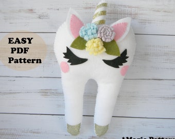 Puede incluir: Un diente de fieltro blanco con cara de unicornio, cuerno de purpurina dorada y una corona de flores de fieltro rosa, azul y amarillo. El texto "EASY PDF Pattern" está en un círculo marrón.