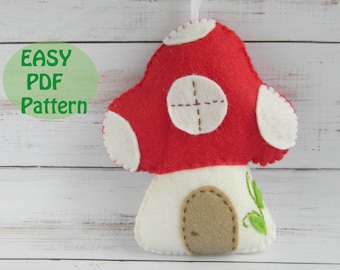 Puede incluir: Adorno de fieltro con forma de hongo, con sombrero rojo y manchas blancas. El hongo tiene una base blanca con una puerta marrón y detalles de hojas verdes. El texto "EASY PDF Pattern" está en un círculo verde.
