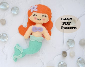 May include: A felt mermaid doll with orange hair, a purple seashell bra, and a teal tail. The doll is smiling and has a flower in her hair. The text "EASY PDF Pattern" is in the background.