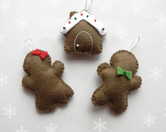 May include: Three felt Christmas ornaments: a gingerbread house with white icing and red and green sprinkles, a gingerbread person with a red bow, and a gingerbread person with a green bow.