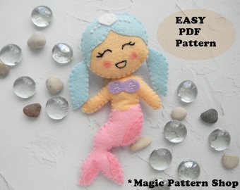 May include: A felt mermaid doll with blue hair, a pink tail, and a purple seashell bra. The doll is holding a white seashell in its hand. The doll is on a white background with small white stones and glass beads.