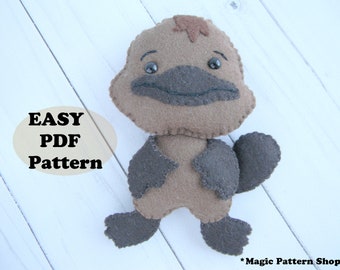 May include: A brown felt plush toy platypus with black felt eyes and a black felt beak. The toy is sitting on a white wooden surface. The text "EASY PDF Pattern" is written on a beige oval shape.