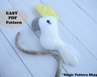 May include: A white and yellow felt cockatoo bird plush toy with black eyes. The toy is sitting on a branch. The text "EASY PDF Pattern" is on a beige oval behind the toy.