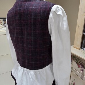 May include: A plaid vest in shades of burgundy, gray, and blue, layered over a white shirt with a ruffled hem. The vest has a classic square pattern and a dark trim.