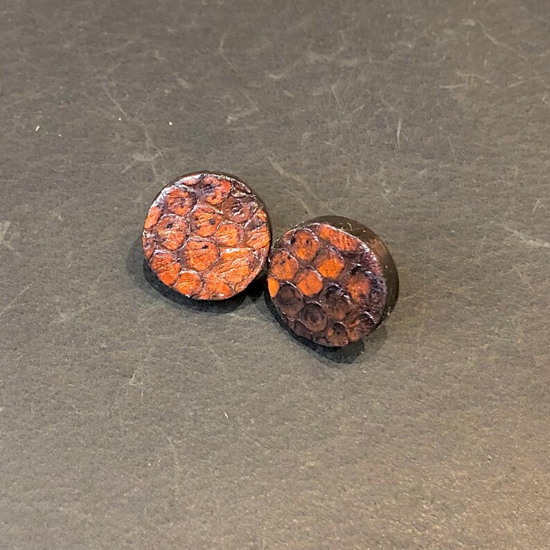May include: Two round wooden plugs with a textured, orange and brown pattern. The plugs have a dark brown border and a flat back. The plugs are likely for ear piercings.