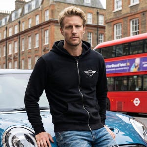 May include: Black zip-up hoodie with the white MINI logo on the chest. The hoodie has a drawstring hood and a full-length zipper. The person is wearing blue jeans and is leaning on a blue car. A red double-decker bus is in the background.