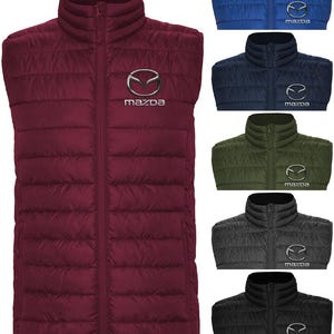May include: A burgundy puffer vest with a full-length zip and a stand-up collar. The Mazda logo is embroidered on the left chest. Additional vests in blue, navy, olive green, charcoal, and black are also shown.