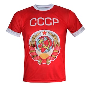 Puede incluir: Camiseta roja con ribete blanco en el cuello y las mangas. La camiseta presenta el texto "CCCP" en letras blancas en la parte superior y un emblema detallado con una hoz y un martillo, un globo terráqueo y espigas de trigo.