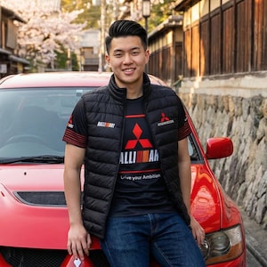 Puede incluir: Un chaleco negro acolchado y una camiseta negra con detalles rojos y el texto "RALLIART" y "Mitsubishi Motors". La camiseta también tiene el texto "Live your Ambition". La persona está apoyada en un coche rojo.