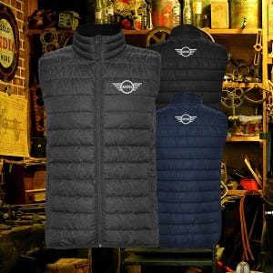 May include: Three quilted vests in black, dark gray, and navy blue. Each vest features a stand-up collar, a full-length zipper, and the "MINI" logo on the chest or back. The vests are displayed against a backdrop of vintage tools and shelves.