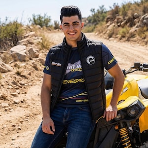 Könnte beinhalten: Eine lächelnde Person trägt eine schwarze Steppweste und ein blaues T-Shirt mit den Logos "Can-Am" und "an-a TEAM". Die Person lehnt an einem gelben ATV. Der Hintergrund ist eine unbefestigte Straße und felsiges Gelände.