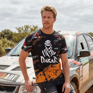 Op de afbeelding: Zwart T-shirt met een Dakar rally-ontwerp. Het shirt heeft een witte afbeelding van een figuur met een hoofdbedekking en het woord "DAKAR" in het wit. Het shirt heeft oranje en witte accenten en de woorden "Ralli Art".