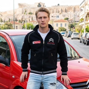 May include: Black zip-up jacket with white trim and the Citroën Racing logo. The jacket has the Total logo on the left chest. The person is standing in front of a red car. The background includes buildings and a historic landmark.