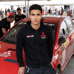 May include: A black zip-up hoodie with the Ralliart logo on the left chest and the Mitsubishi Motors logo on the right. The hoodie is worn by a person standing in front of a red Mitsubishi rally car.