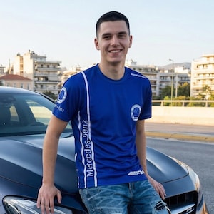 Könnte beinhalten: Ein blaues Kurzarm-T-Shirt mit weißen Streifen und dem Text "Mercedes-Benz" an der Seite. Das Shirt hat ein Logo auf der Brust und das AMG-Logo auf dem Ärmel. Die Person trägt eine blaue Jeans.